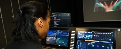 Young woman in a dark room sitting facing a bank of screen showing data